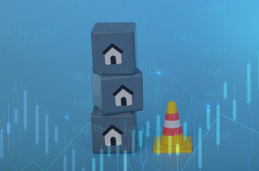 Three blocks with pictures of housing on them stacked precariusly, with a traffic cone next to them