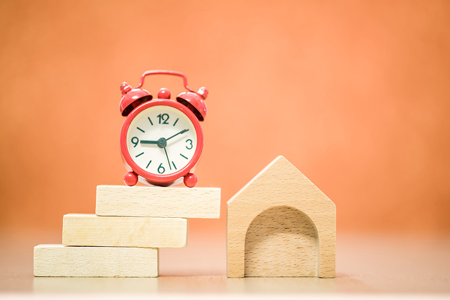 A red alarm clock on a pile of wooden bricks next to a wooden house