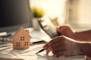 A close up of a hand holding a pen, next to a small wooden house and a calculator