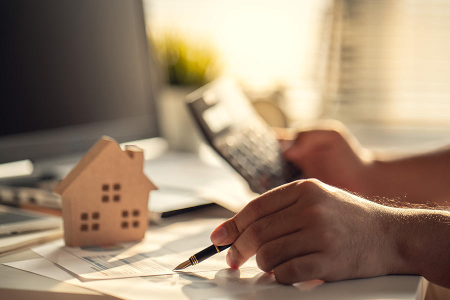 A close up of a hand holding a pen, next to a small wooden house and a calculator