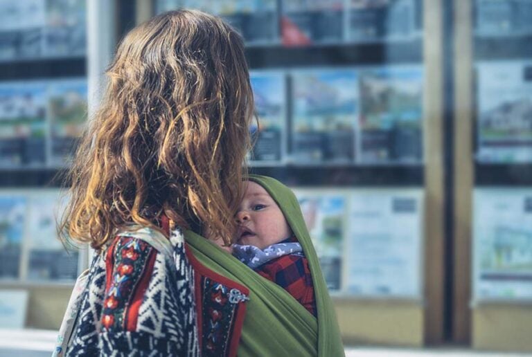 A young woman with a baby in a sling looks in an estate agent's window