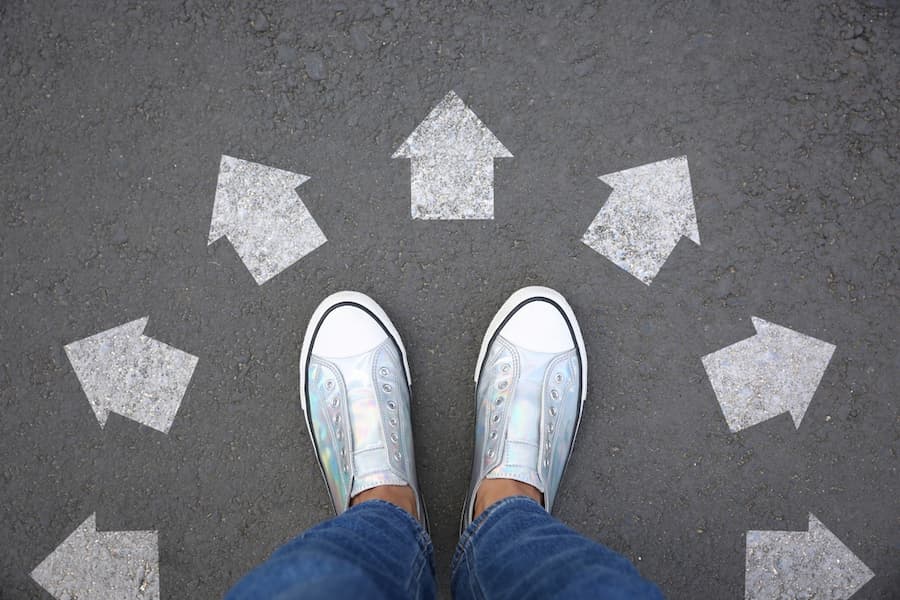Woman standing near white arrows pointing in different directions on asphalt road, top view. Concept of choice and making decisions