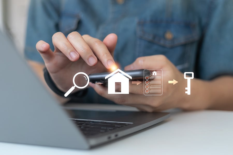 A close-up of hands holding a phone over a laptop, with an overlaid graphic of a house, document and key