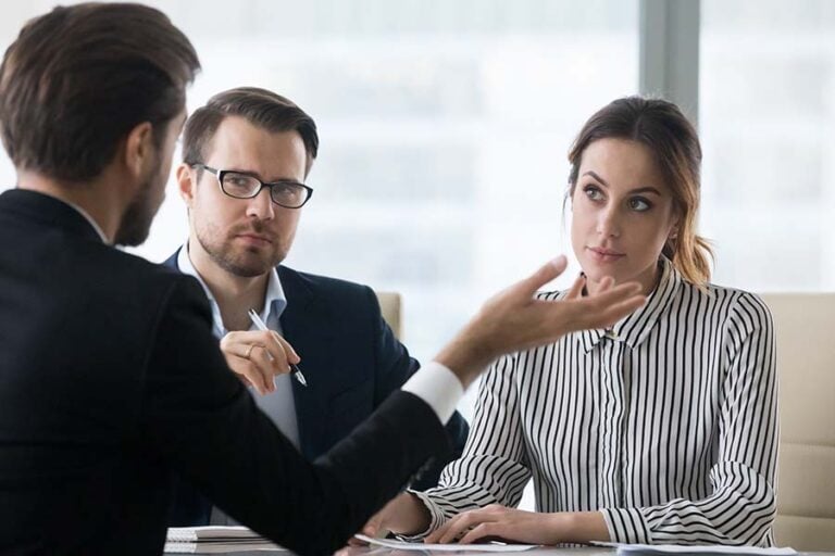 A lawyer seen from behind dressed in black suit gesticulates in front of a suspicious looking couple