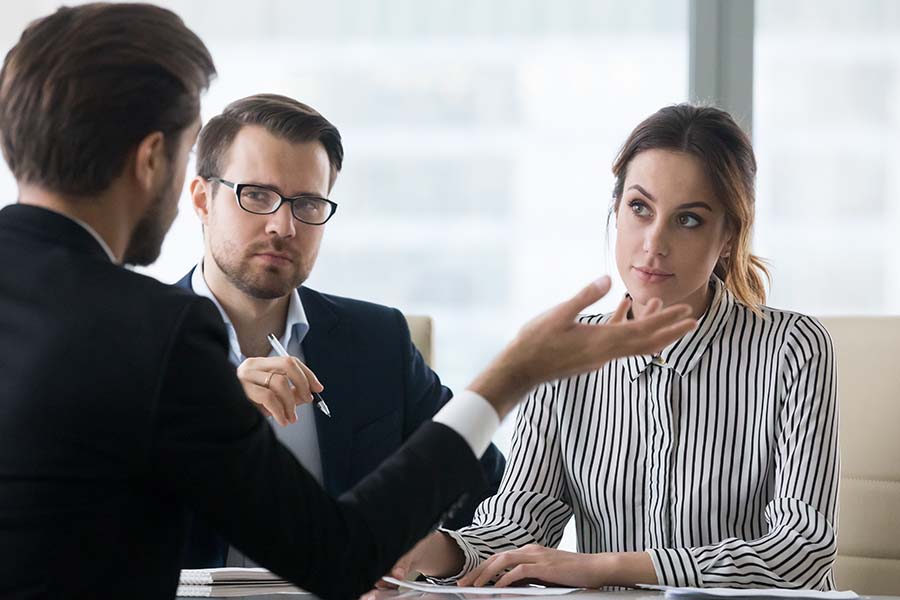 A lawyer seen from behind dressed in black suit gesticulates in front of a suspicious looking couple