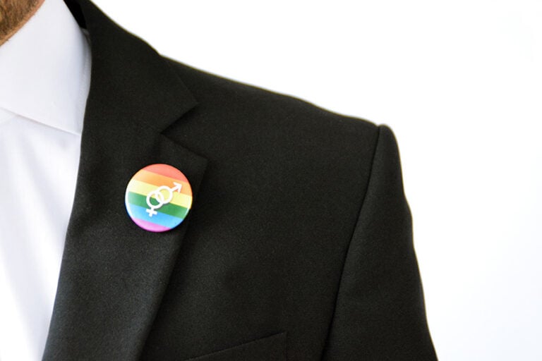A close-up of a man wearing an LGBTQ pin in rainbow colours on a black suit lapel
