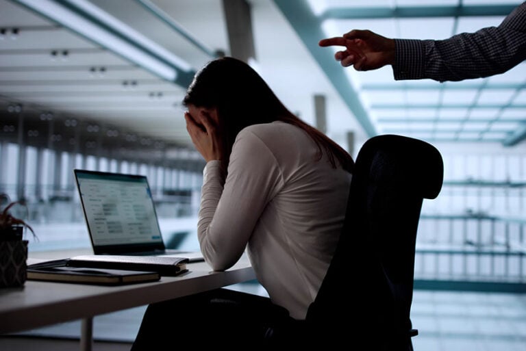 A bullied woman at her desk in an office being pointed at by an angry man
