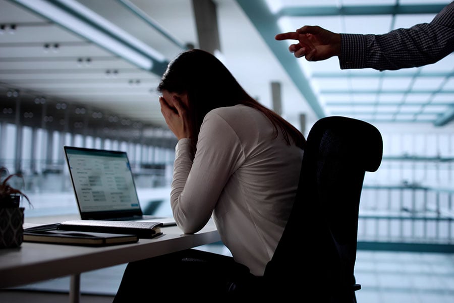 A bullied woman at her desk in an office being pointed at by an angry man