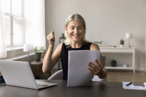 A middle-aged woman looks happy as she reads from papers she's holding, while sitting at a table with a laptop
