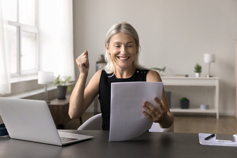 A middle-aged woman looks happy as she reads from papers she's holding, while sitting at a table with a laptop