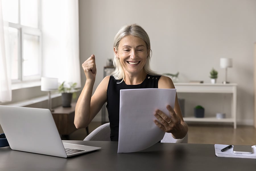 A middle-aged woman looks happy as she reads from papers she's holding, while sitting at a table with a laptop