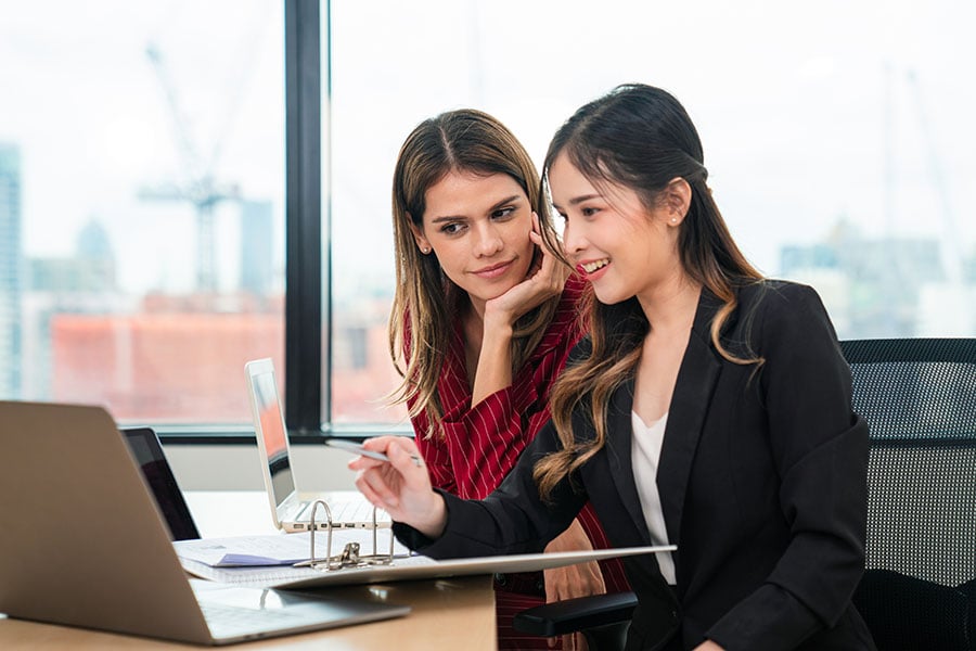 A senior employee supervises a younger employee at a desk containing two laptops and files