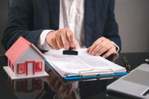 An estate agent stamps a pile of papers on a desk, next to a small model house