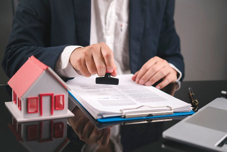 An estate agent stamps a pile of papers on a desk, next to a small model house