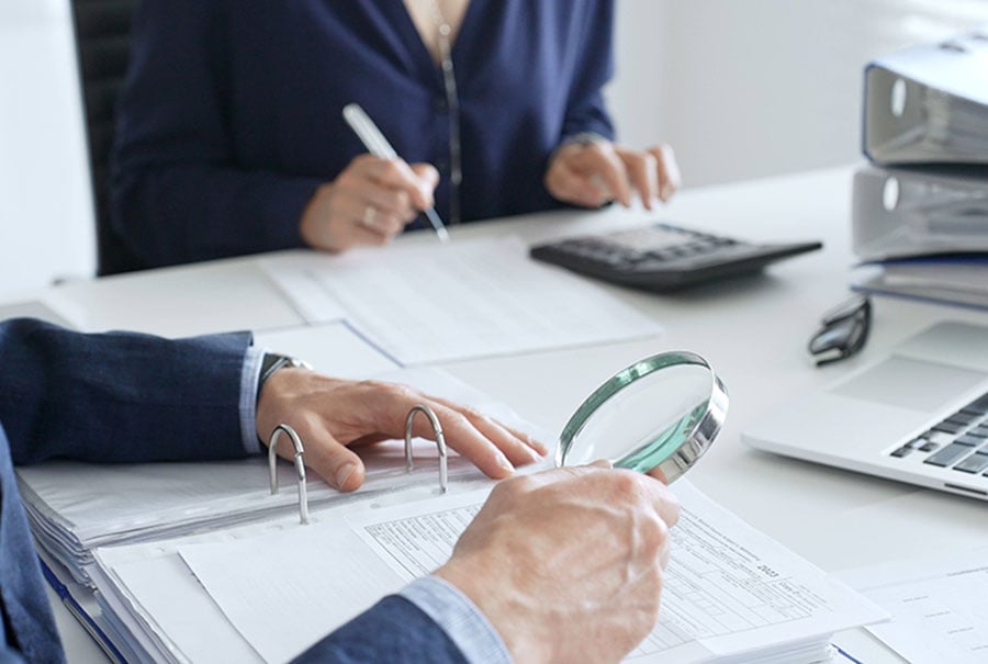 Two professionals at a desk inspecting a pile of files, using a magnifying glass and a calculator