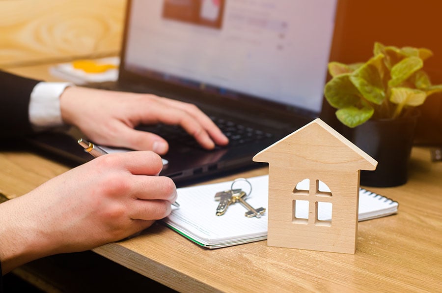 Close-up of hands writing on a laptop keyboard, next to a notepad and a small wooden house