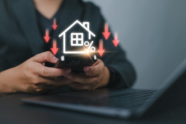 A graphic of a house with red downwards arrows is overlaid on a person's hands holding a calculator