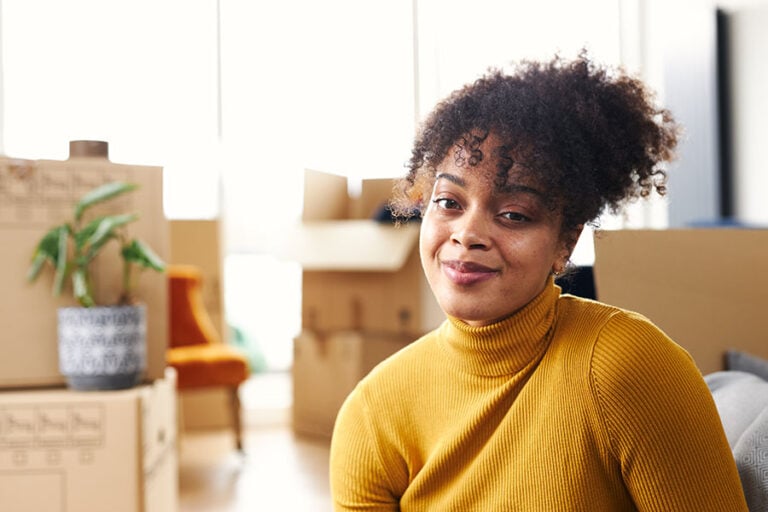 A smiling young woman sits in on the floor in her house surrounded by open packing boxes