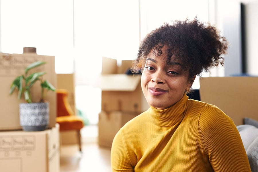 A smiling young woman sits in on the floor in her house surrounded by open packing boxes