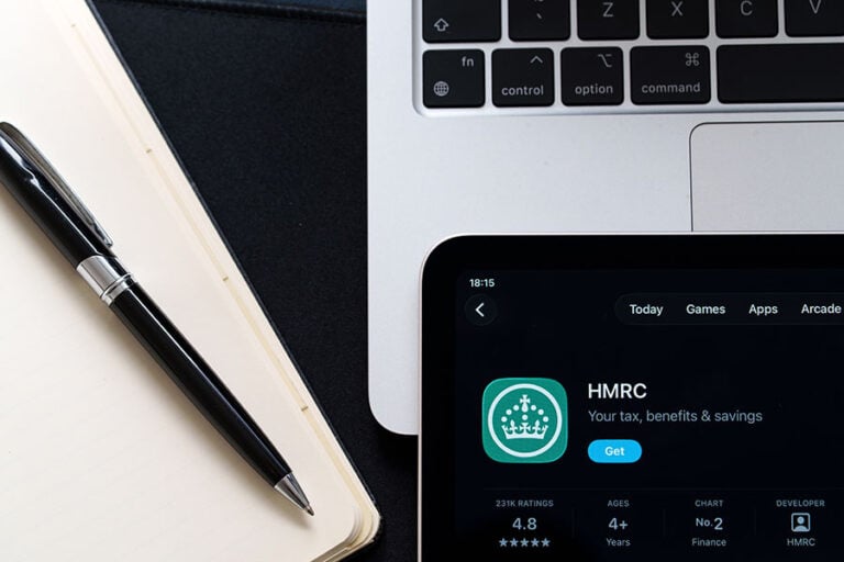 A phone with an HMRC app, laid on top of a laptop and next to a notepad and pen
