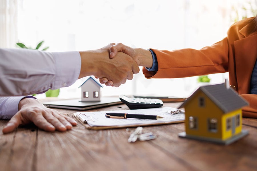 Two people shake hands over a desk with a small house and set of keys on it
