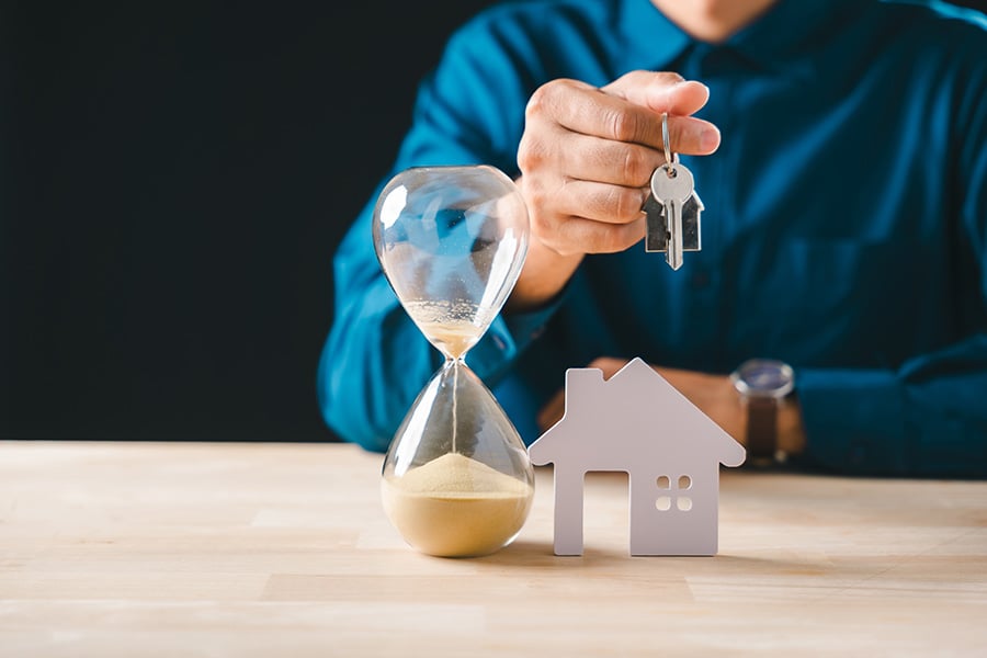A man sits at a desk holding a house key, with a small wooden house in front of him next to an hourglass
