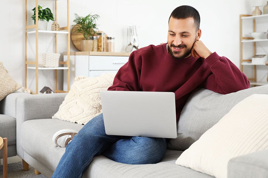 A smiling man using a laptop while sitting on a sofa
