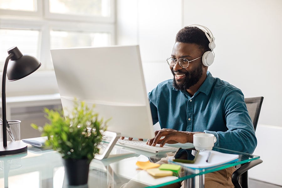 A smiling man wearing headphones sits at a desk in front of a computer taking notes
