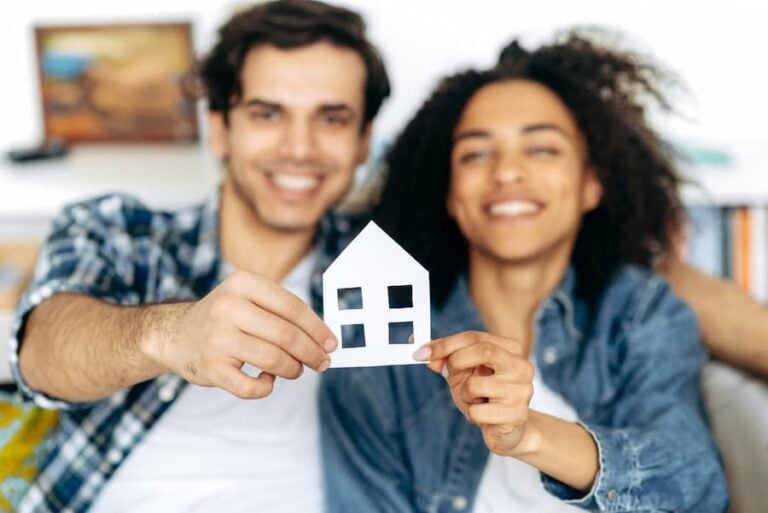 Joyful multiethnic couple holding paper house, sitting on a sofa in their new modern apartment